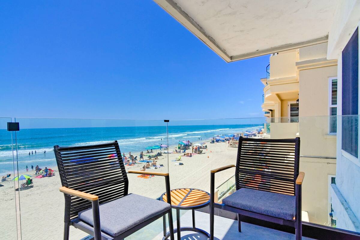 A balcony with two chairs and a small table overlooking a beach with people and umbrellas.
