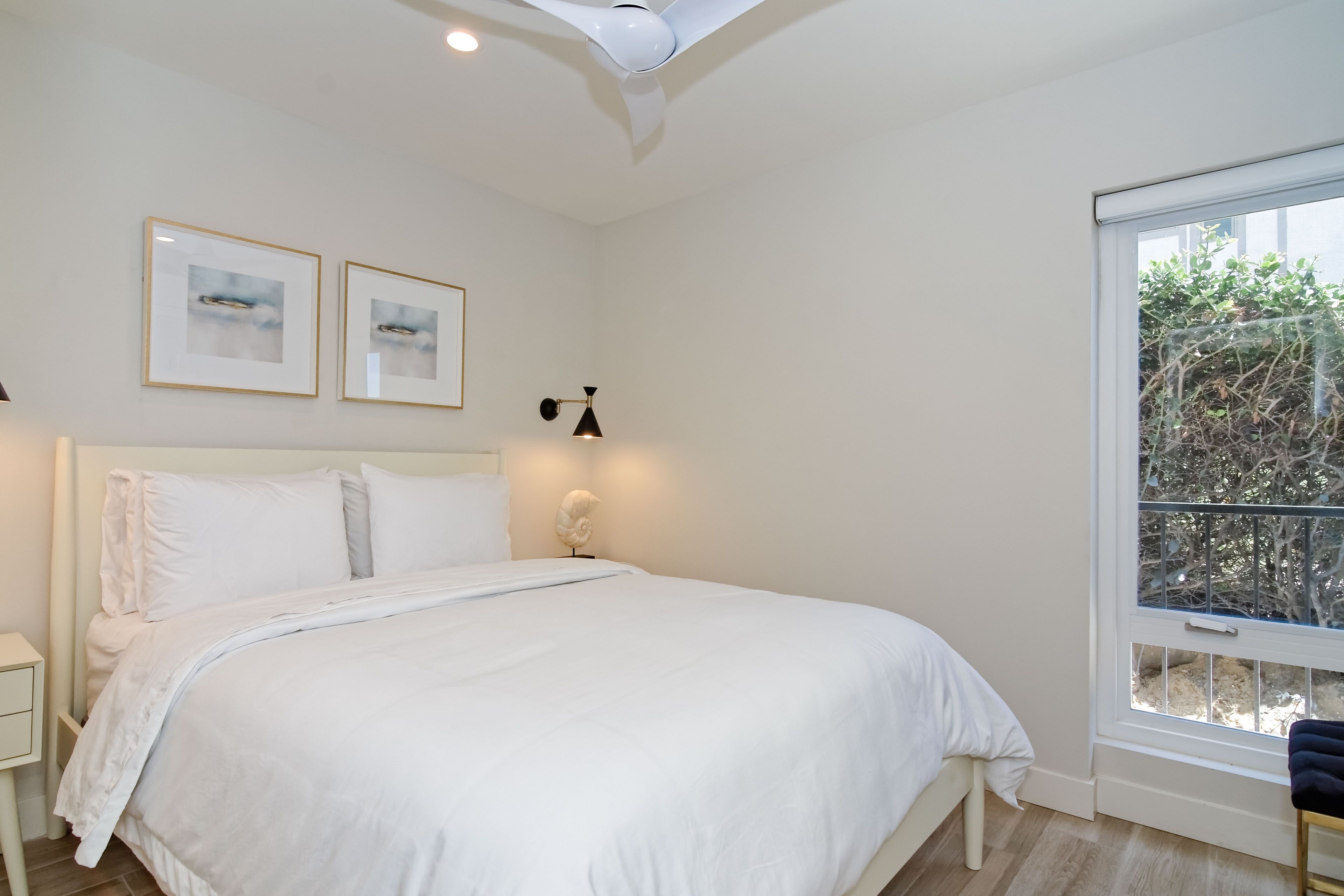 Quarto com uma cama grande, paredes brancas, duas molduras com quadros, abajur e janela com vista para o verde.