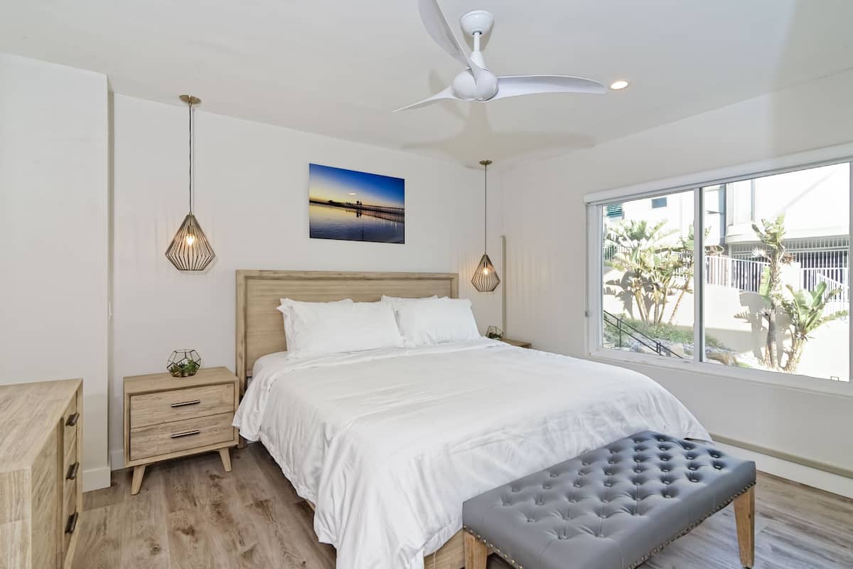 A bedroom with a large bed, a ceiling fan, a nightstand, a bench, and a window with a view of palm trees.