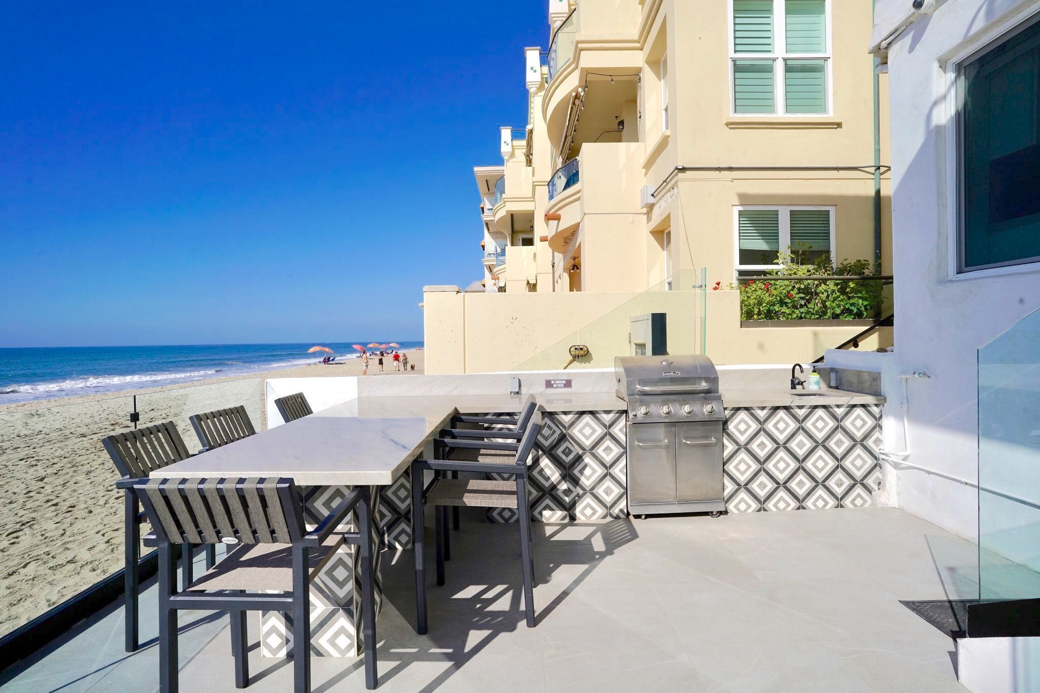 A beachside patio with a table and chairs, a grill, and a view of the ocean.