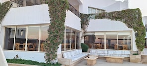 Un espace de détente extérieur moderne, avec un canapé blanc et des bancs en bois, entouré de grandes fenêtres et d’un patio couvert.