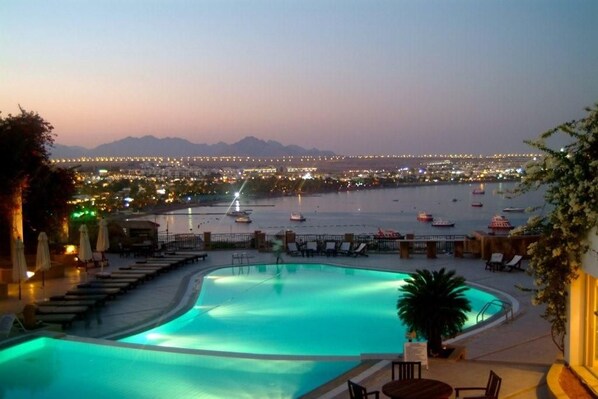 A swimming pool with lounge chairs and umbrellas, overlooking a marina with boats and city lights at dusk. - Eden Rock Hotel - Sharm El Sheikh (Sharm El Sheikh)