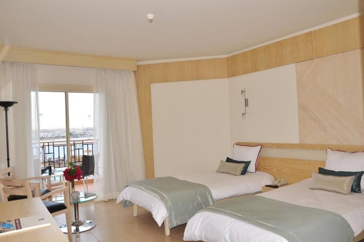 A hotel room with two beds, a balcony with a view, a dining area, and a small table with a vase of flowers.