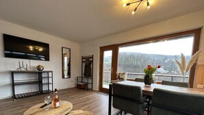 A modern living room with a wooden floor, a dining table with chairs, a sofa, a TV mounted on the wall, and a large window with a view of trees and hills. - FRITZ - Haus Sonnenblick (Diemelsee)