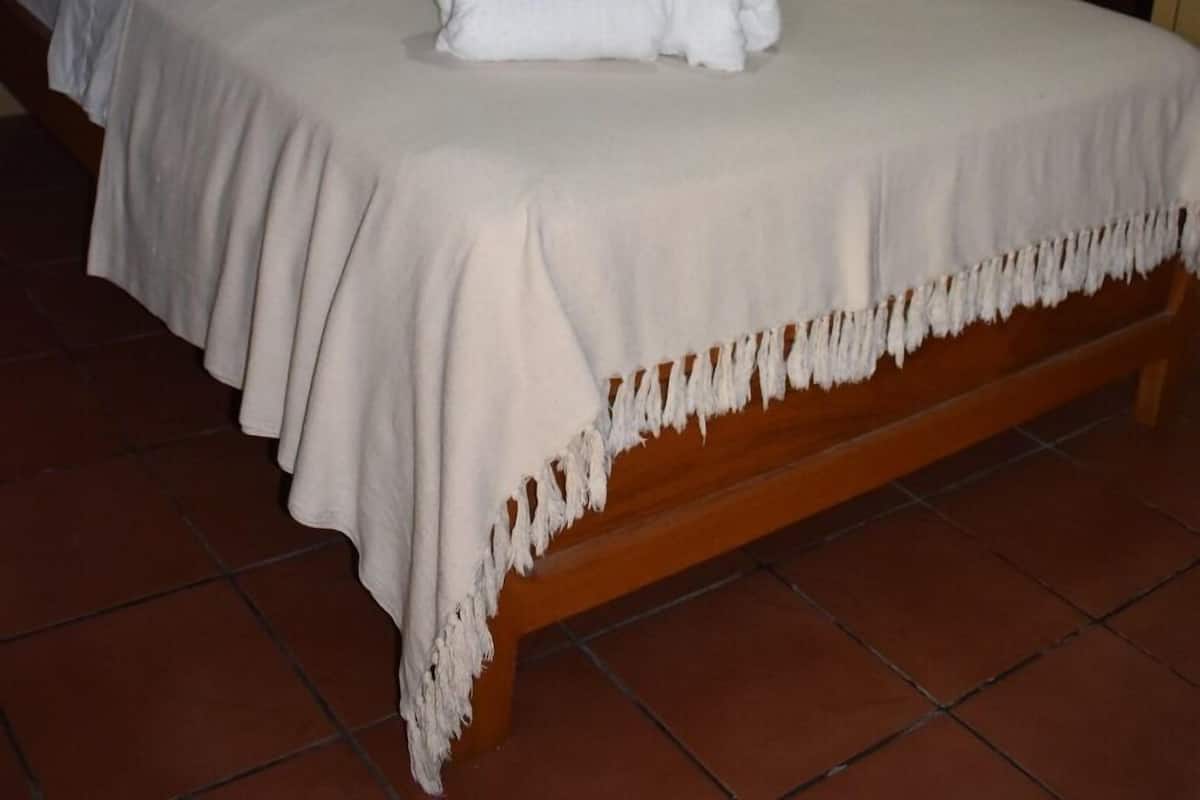 Una cama bien hecha con una colcha blanca y una toalla, un cabecero de madera, pared de ladrillo y un equipo de aire acondicionado sobre la cama.