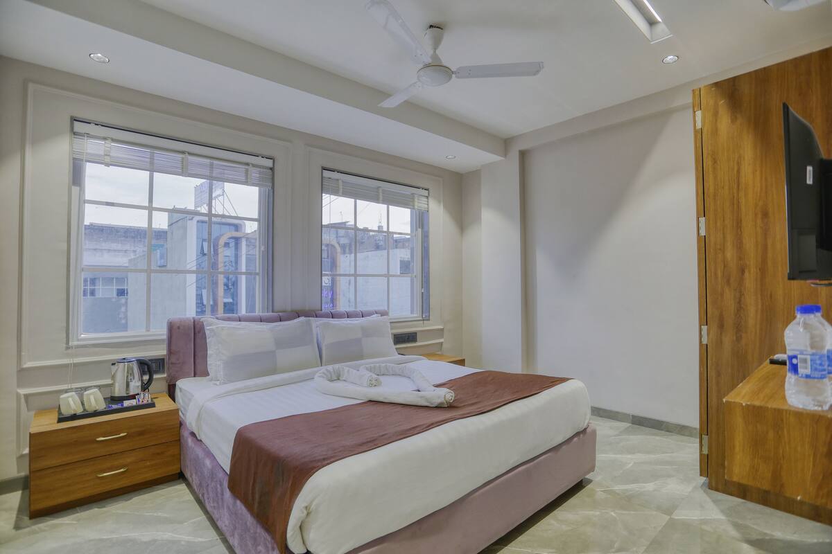 Habitación de hotel con cama, mesita de noche con teléfono y tetera, armario de madera y ventana grande con persianas.