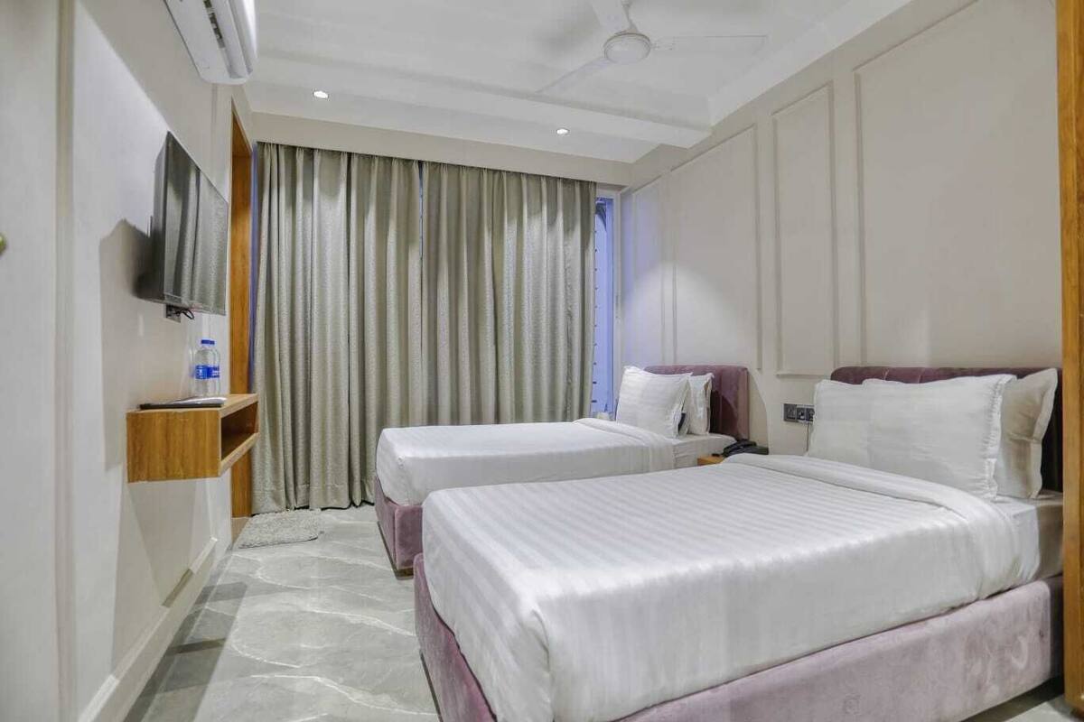 Habitación de hotel con dos camas, un televisor montado en la pared, una botella de agua sobre una mesita auxiliar y una ventana con cortinas.
