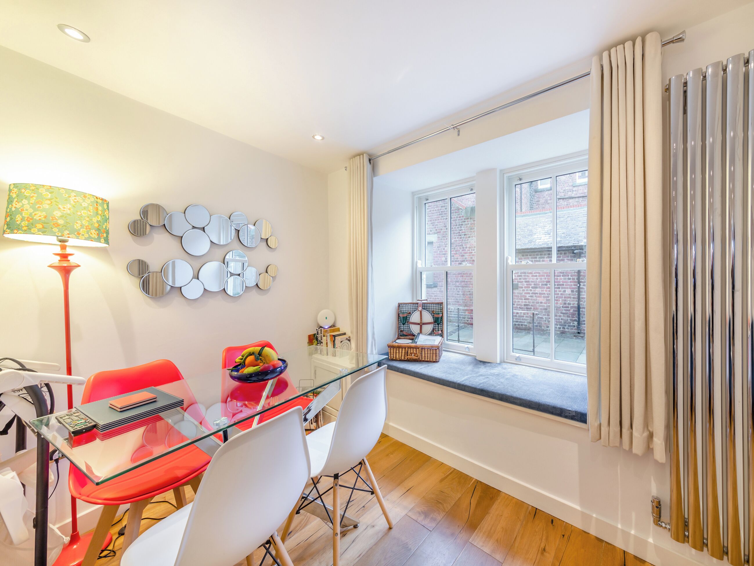 Un espace repas moderne avec une table en verre, des chaises rouges, une lampe et une vue sur un bâtiment en briques, visible à travers une fenêtre.