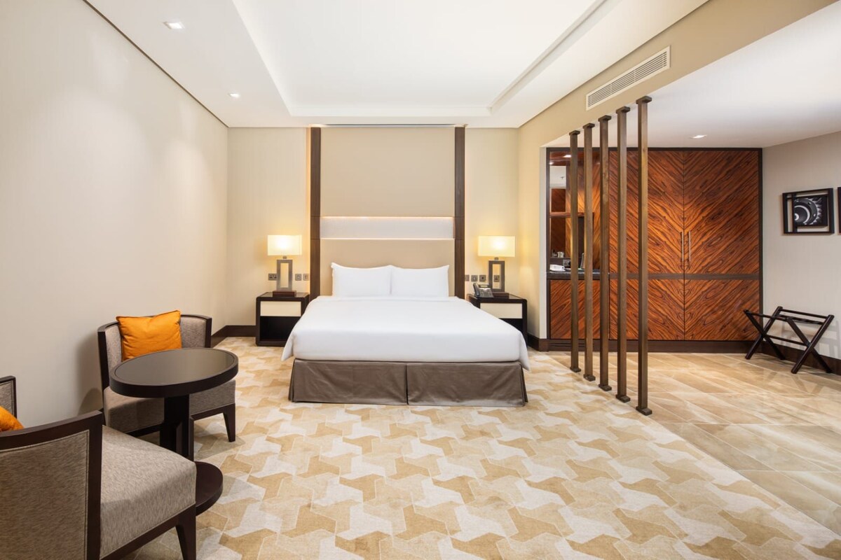 Una habitación de hotel con cama, mesitas de noche, una silla, una pequeña mesa y un panel decorativo en la pared.