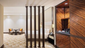 Une chambre d’hôtel avec un lit, un bureau avec un téléphone, une machine à café et une petite table avec des chaises. Un panneau décoratif en bois orne le mur, et un tapis à motifs recouvre le sol.