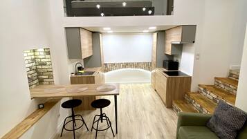 A modern kitchen with a central island, a dining area with stools, a sofa, and a staircase leading to an upper level.