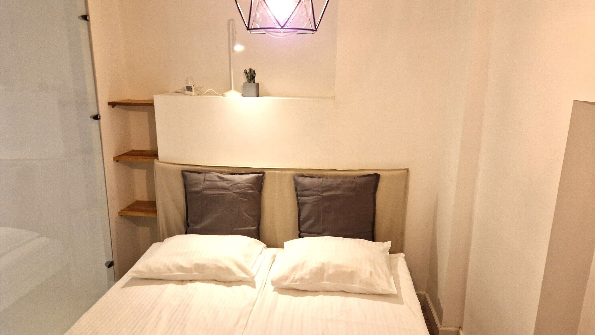 Una camera da letto con un letto, due cuscini, un comodino con una lampada e una mensola con una pianta.
