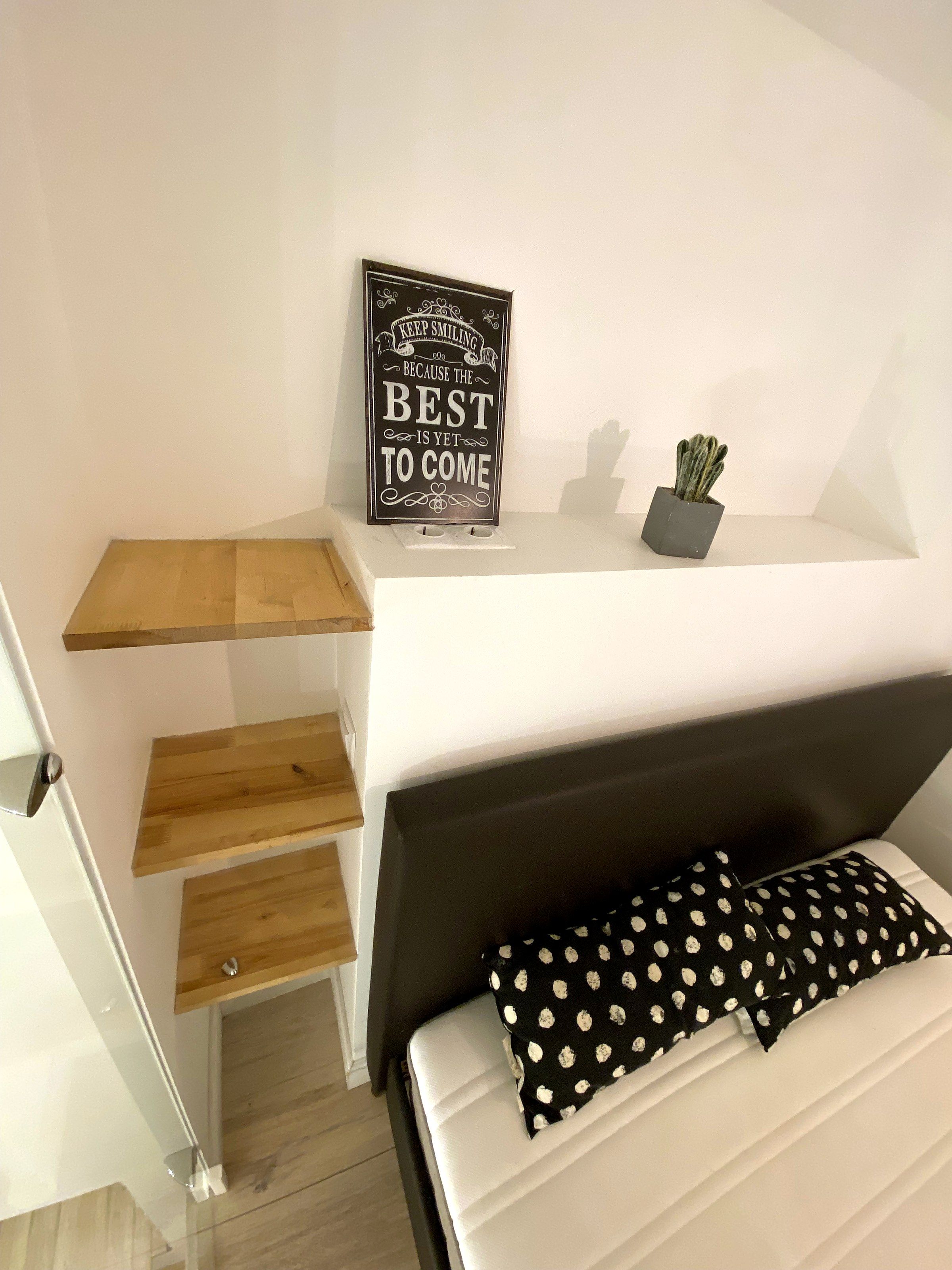 A bedroom with a bed, a bedside table, a wall-mounted shelf with a motivational sign, and a small potted plant.