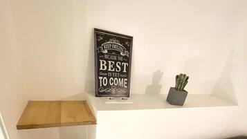 A bedroom with a bed, a bedside table, a wall-mounted shelf with a motivational sign, and a small potted plant.