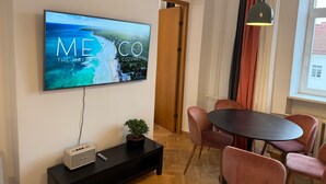 Una sala moderna con un televisor de pantalla plana mostrando 'MEXICO THE MEXICAN RIVER & COZUMEL', una puerta de madera, un área de comedor con sillas rosadas y una mesa negra.