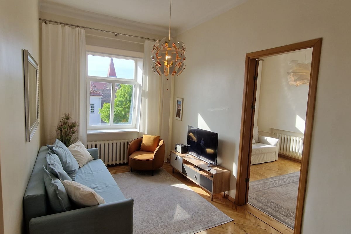 A living room with a sofa, a TV, a chair, and a window with a view of a building and trees.