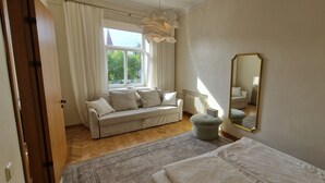 Un dormitorio con cama, sofá, un pequeño otoman, un espejo grande y una ventana con cortinas.