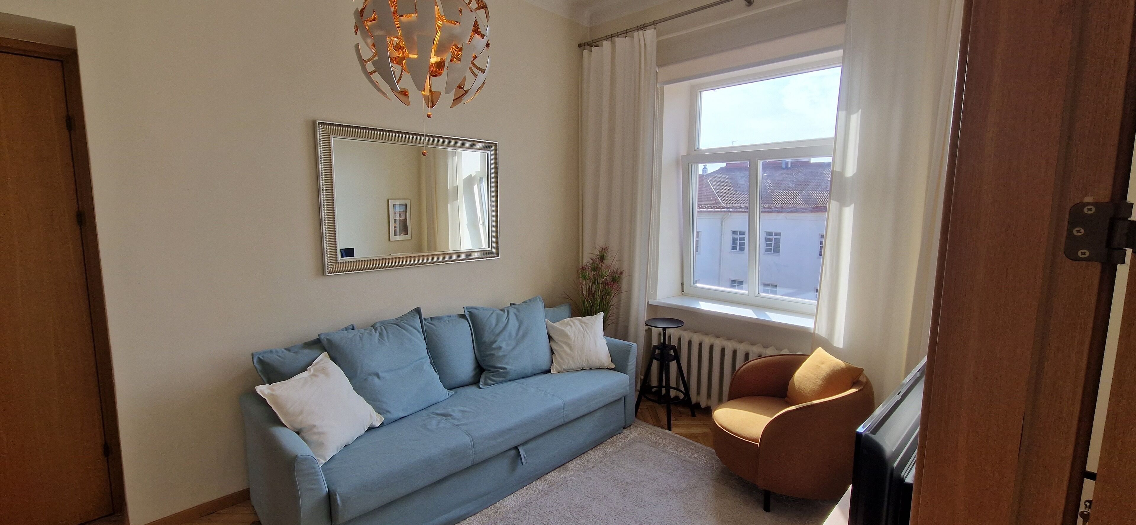 Un salon avec un canapé bleu, un fauteuil marron, un grand miroir et une fenêtre donnant sur des immeubles.