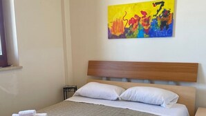 A neatly made bed with white linens and rolled towels, a wooden headboard, and a colorful abstract painting on the wall.