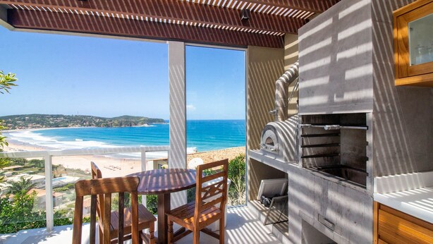 Un balcon avec vue sur la plage et l’océan, une table et des chaises en bois, et un barbecue encastré.