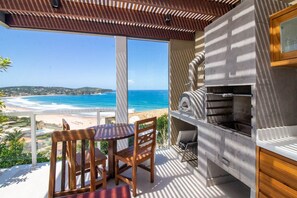 A balcony with a view of the beach and ocean, a wooden table and chairs, and a built-in grill. - Vista Panorâmica em Geribá com 5 suítes (Búzios)