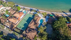 An aerial view of a coastal resort with a tennis court, swimming pool, and beach access. - Cinematográfica Mansão Pé na Areia  (Búzios)