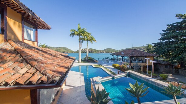 A swimming pool with a view of the sea, surrounded by palm trees and a building with a tiled roof. - Cinematográfica Mansão Pé na Areia  (Búzios)