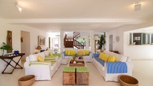 A spacious living area with a central coffee table, two sofas with striped and solid cushions, a wooden staircase, and a large window providing natural light. - Harmoniosa Mansão com 7 Suítes Ferradura (Búzios)