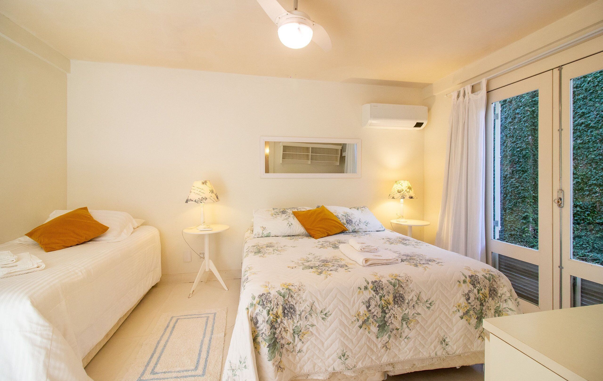 Un dormitorio con dos camas, un ventilador de techo, una ventana con cortinas y una pequeña mesa con lámpara.