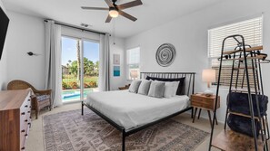 A bedroom with a bed, a ceiling fan, a nightstand, a lamp, a chair, a window with blinds, and a view of a swimming pool. - Escape the Cold - Fall & Winter Rates Reduced! (Indio)