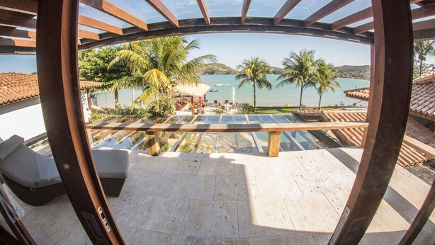 Vista panorámica desde un espacio al aire libre cubierto con una pérgola de madera, con vista a una playa con palmeras y un mar azul cristalino.