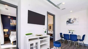 A modern hotel room with a white console table, a flat-screen TV mounted on the wall, a dining area with blue chairs, and a large window. - Muse:5 minutes to broadway

 (Nashville)