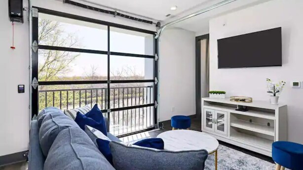 A modern living room with a large sliding glass door, a sofa with blue cushions, a round coffee table, a white cabinet, and a mounted TV. - Muse:5 minutes to broadway

 (Nashville)