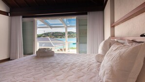 A bedroom with a large bed, white linens, and a view of a coastal area with buildings and a blue sky. - Mansão Pé na Areia Ferradura com 4Suítes (Búzios)