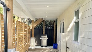 A covered outdoor area with a grill, wooden fence, and a blue trash bin.