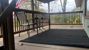 A covered outdoor deck with a black mat, two chairs, a table, and an American flag. - Home away from home (Waldron)