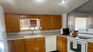 A compact kitchen with wooden cabinets, a white sink, a dishwasher, a stove, and a window with blinds. - Home away from home (Waldron)