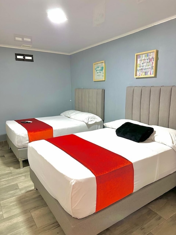 Habitación de hotel con dos camas, cada una con ropa blanca y un detalle rojo. Hay dos cuadros en la pared, y la habitación tiene paredes azul claro y suelo de madera.