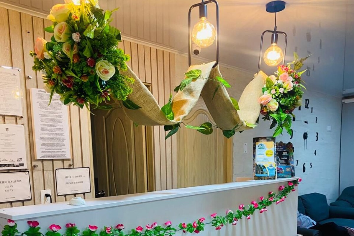 Uma mesa de recepção com superfície branca, flores artificiais verdes e um relógio na parede.