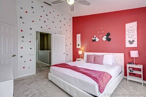 A bedroom with a white bed, red walls, Mickey Mouse decorations, and a ceiling fan.