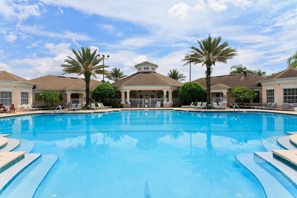A swimming pool with lounge chairs and palm trees, surrounded by buildings with a central pavilion.