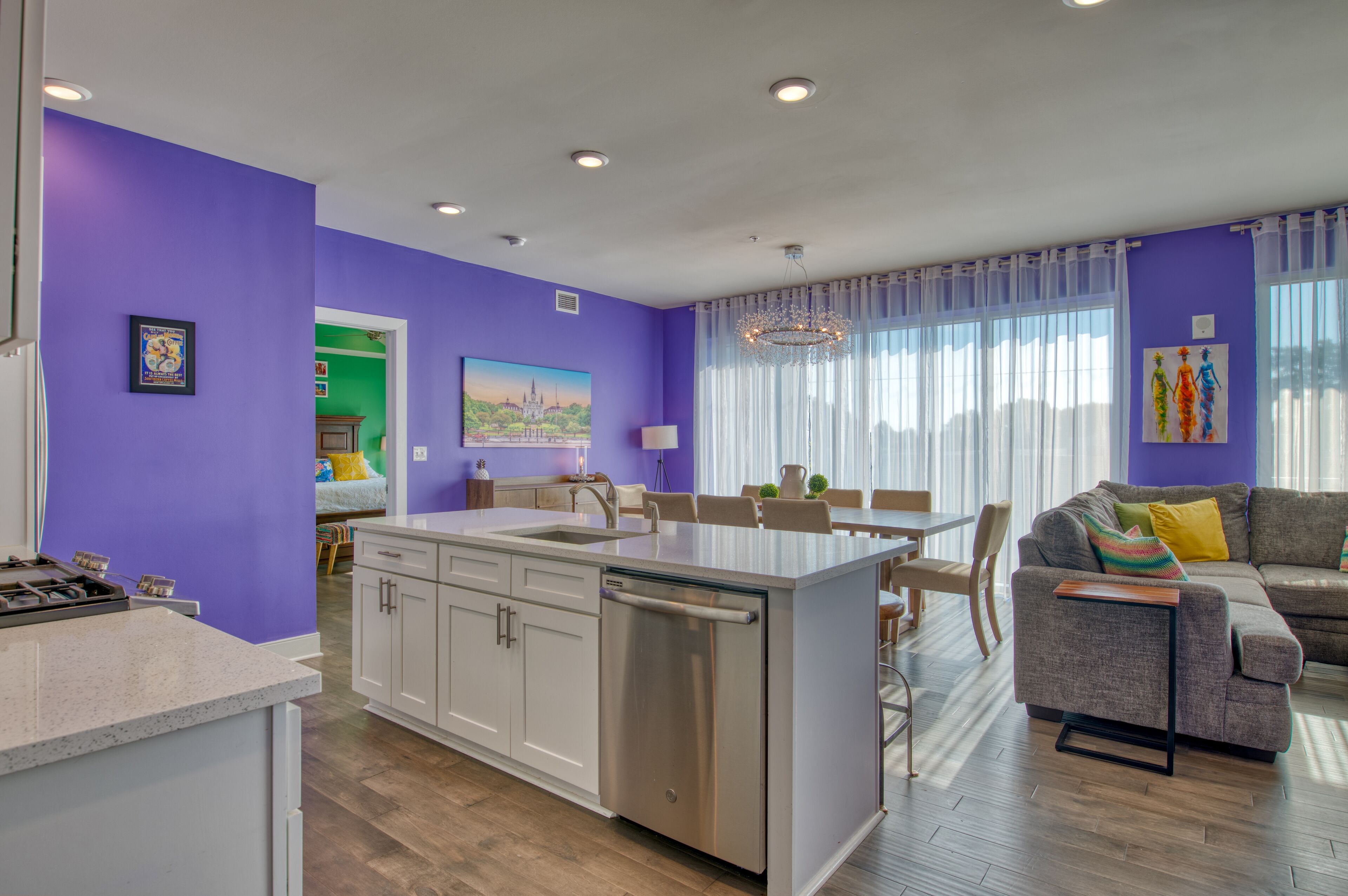 A modern kitchen with white cabinets and a stainless steel dishwasher, a dining area with a table and chairs, and a living room with a sofa and a TV.