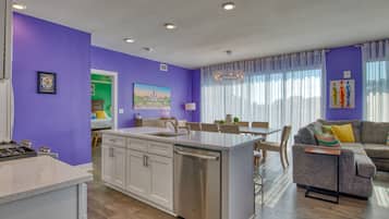 A modern kitchen with white cabinets and a stainless steel dishwasher, a dining area with a table and chairs, and a living room with a sofa and a TV.