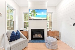 A living room with a fireplace, a television mounted above it, and a view of a beach through the window.