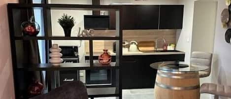 A modern living room with a dark grey armchair, a wooden barrel, and a kitchen area with black cabinets and a wooden countertop.