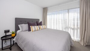 A modern bedroom with a large bed, a bedside table with a lamp, and a window with sheer curtains. - Luxury 3 Bedroom Wanaka Townhouse (Albert Town)