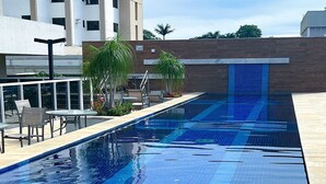 A swimming pool with a tiled floor, surrounded by outdoor seating and a building in the background. - Apto Moderno com Vista Fantástica do Flamboyant (Goiânia)