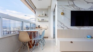 A modern kitchen with a large window, a dining area with a table and chairs, and a TV mounted on a marble wall. - Apto Moderno com Vista Fantástica do Flamboyant (Goiânia)