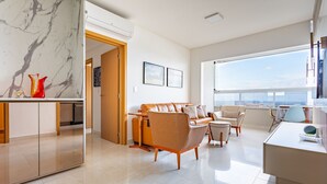 A modern living room with a sofa, chairs, and a dining area. There is a large window with a view of the city. - Apto Moderno com Vista Fantástica do Flamboyant (Goiânia)