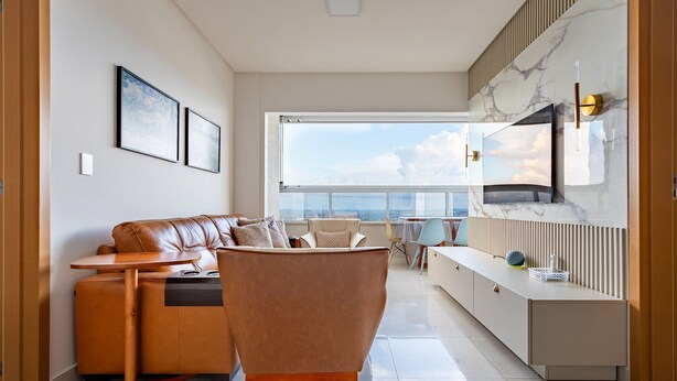 A modern living room with a leather sofa, a wooden coffee table, and a TV mounted on a marble wall. - Apto Moderno com Vista Fantástica do Flamboyant (Goiânia)